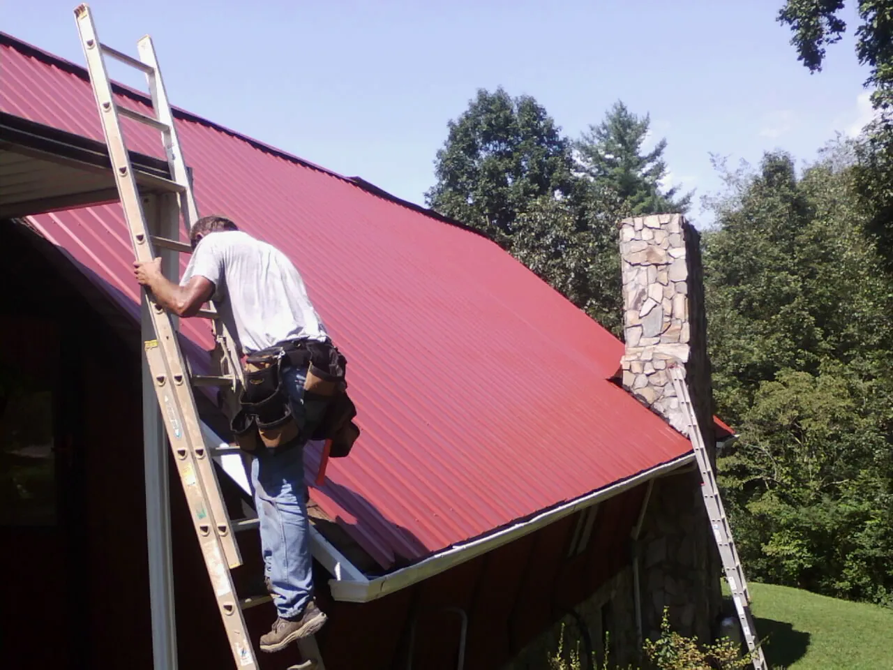Roofer on a ladder beside a red metal roof and stone chimney — Soffit & Fascia Repair by Roofing Brothers in Springdale